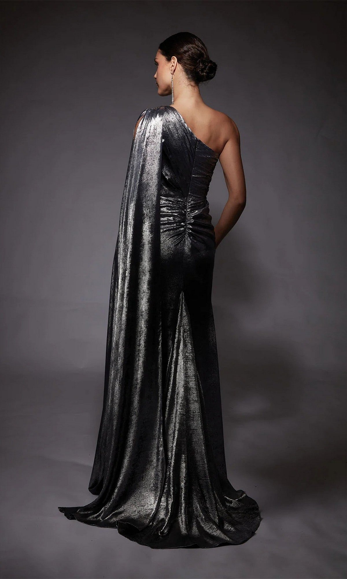A woman stands with her back to the camera in the Alyce 27753 long formal dress, a one-shoulder metallic chiffon gown featuring a draped train, set against a dark background.
